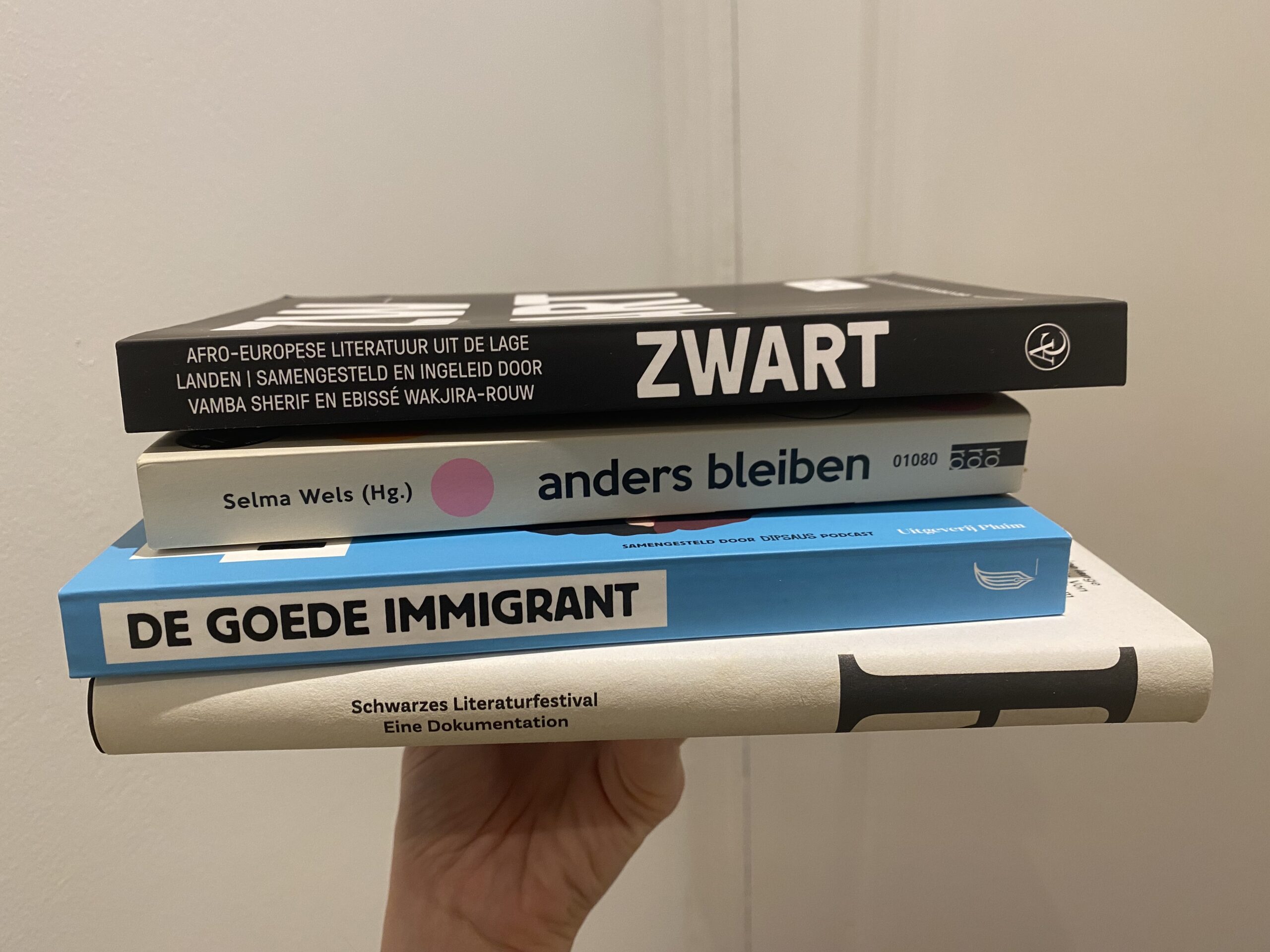 A hand holding up the books "Zwart: Afro-Europese Literatur uit de Lage Landen", "anders bleiben", "De Goede Immigrant", and "Resonanzen: Schwarzes Literaturfestival - Eine Dokumentation"